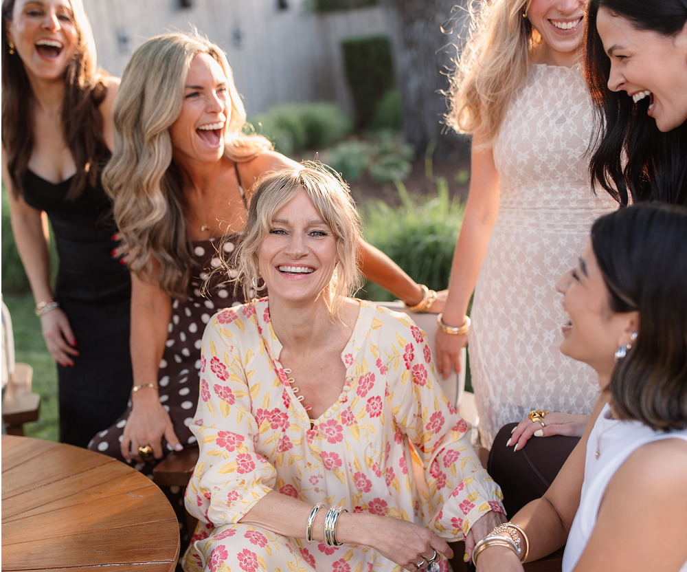 Group of women laughing and smiling outdoors, one seated in floral dress.