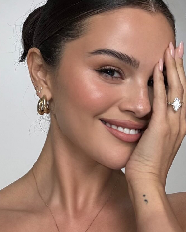 A smiling woman with her hair pulled back holds her hand to her face, showing her jewelry.