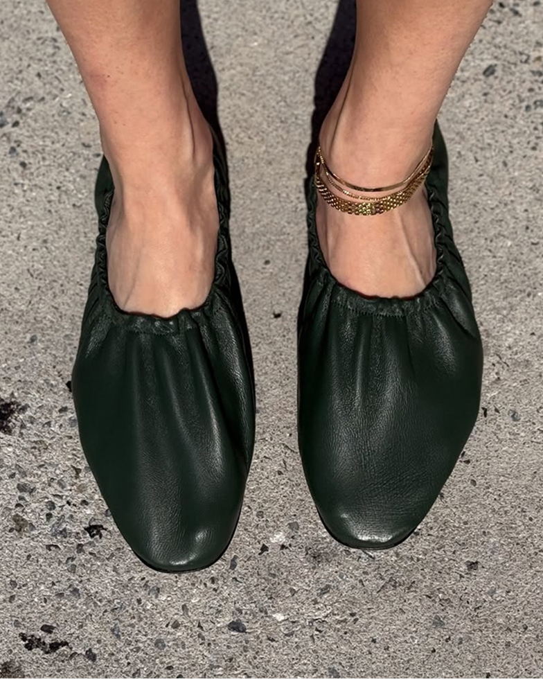 A close-up of a person's feet wearing dark green ruched flats and a gold anklet on concrete pavement.