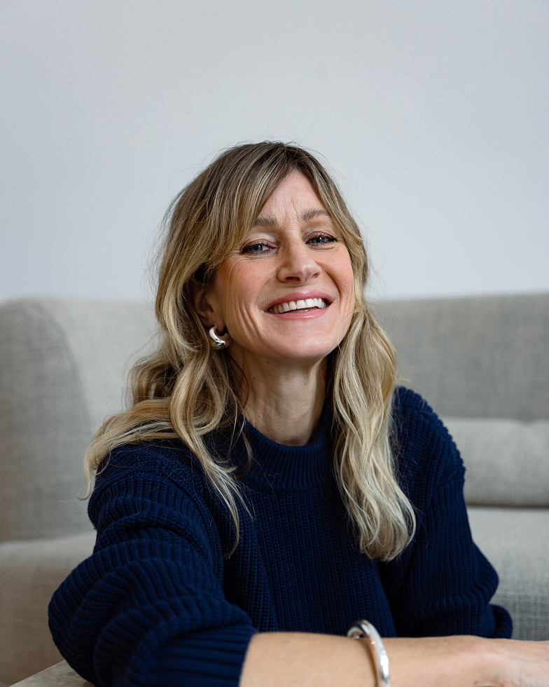 A smiling woman with blonde, wavy hair in a navy blue knit sweater looks at the camera.