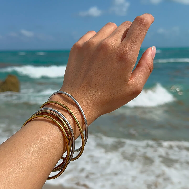 A person's hand wearing several gold and silver bangles with the ocean in the background.