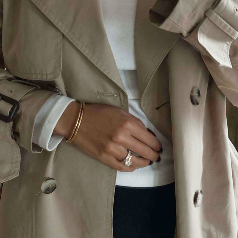 A close-up of a person wearing a beige trench coat, a gold bracelet, and a diamond ring.