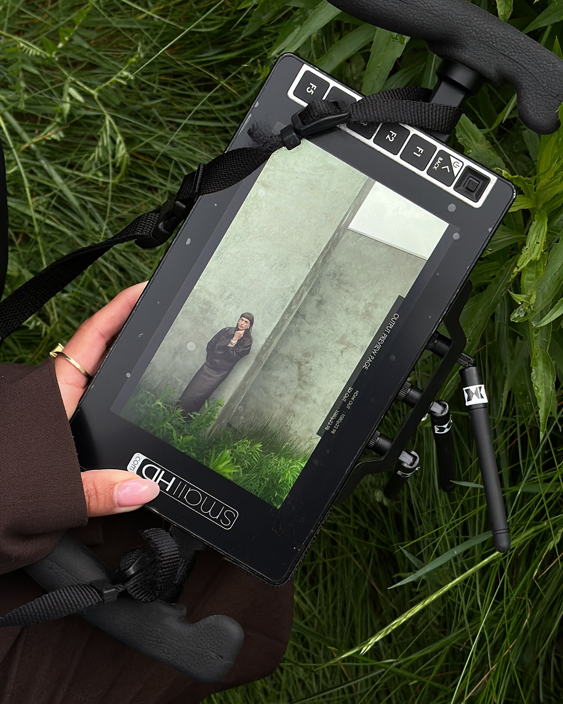 A hand holding a monitor displaying a person standing against a wall.