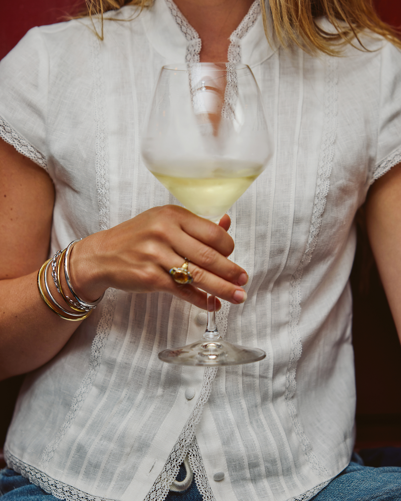 A woman in a white lacy blouse holds a glass of white wine, wearing bangles and a ring.