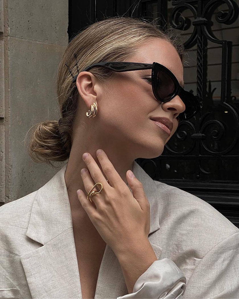 Woman wearing sunglasses, gold earrings, and a beige blazer, with her hair in a low bun.