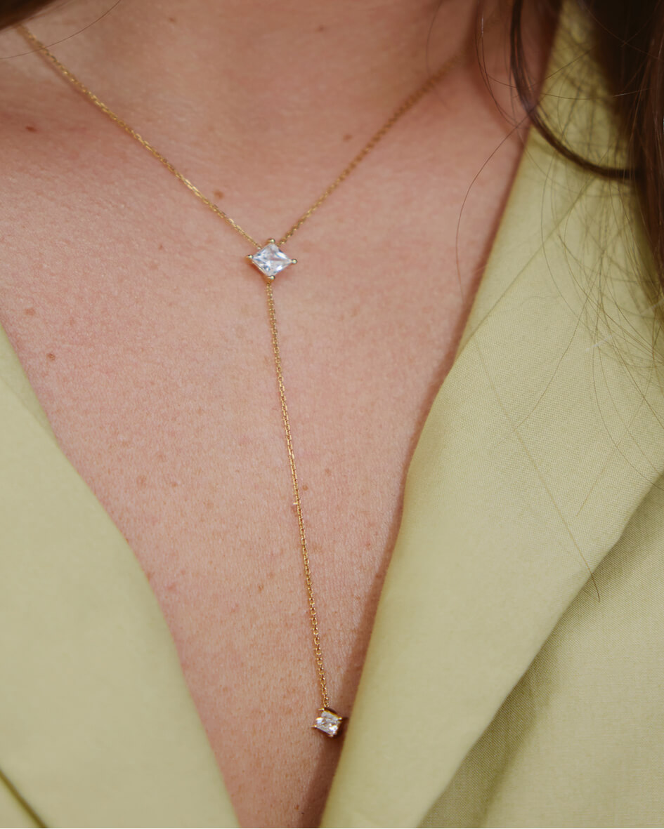 A close-up of a person's neck, wearing a delicate gold lariat necklace with two square-cut stones.