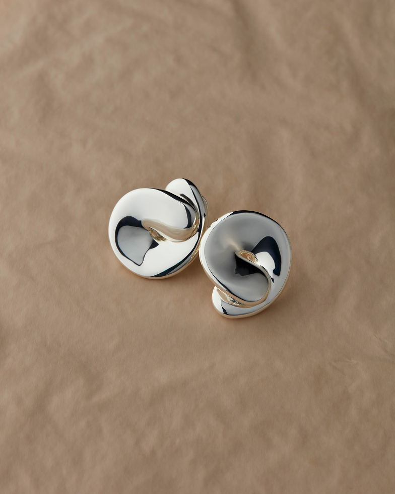 A pair of sculptural, highly polished silver earrings on a smooth, tan surface.