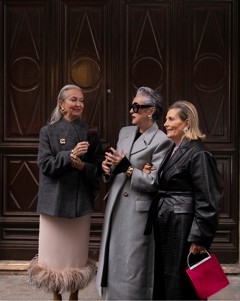 Three stylish older women in coats and jewelry talking in front of a large wooden door.