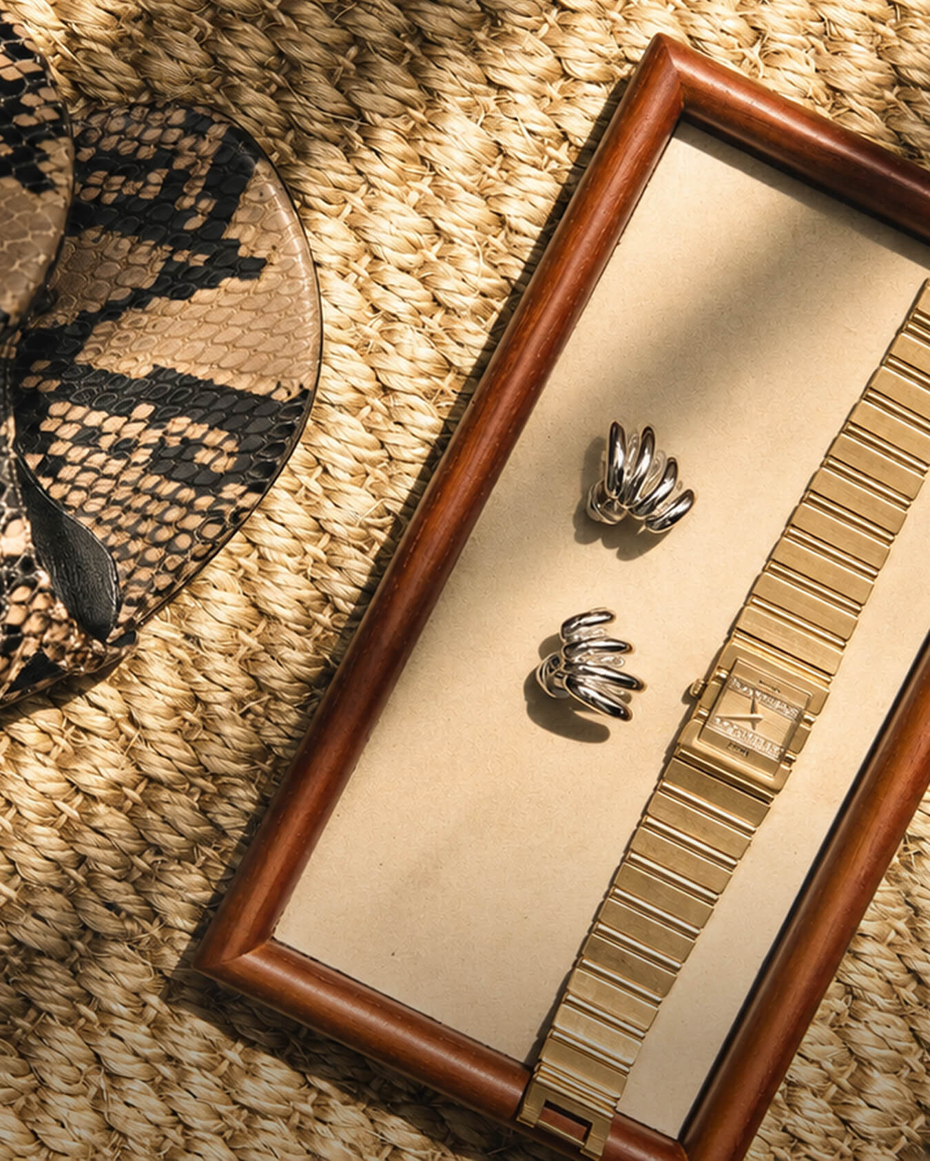 A flat lay of a gold watch, silver earrings, and a snakeskin shoe on a woven surface.