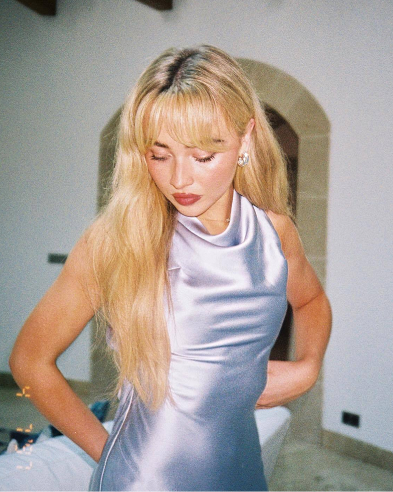 A woman with long blonde hair and bangs in a shiny lavender halter dress looks down.