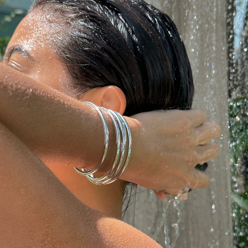 A close-up of a person wearing silver bangles showering and washing their wet, dark hair.