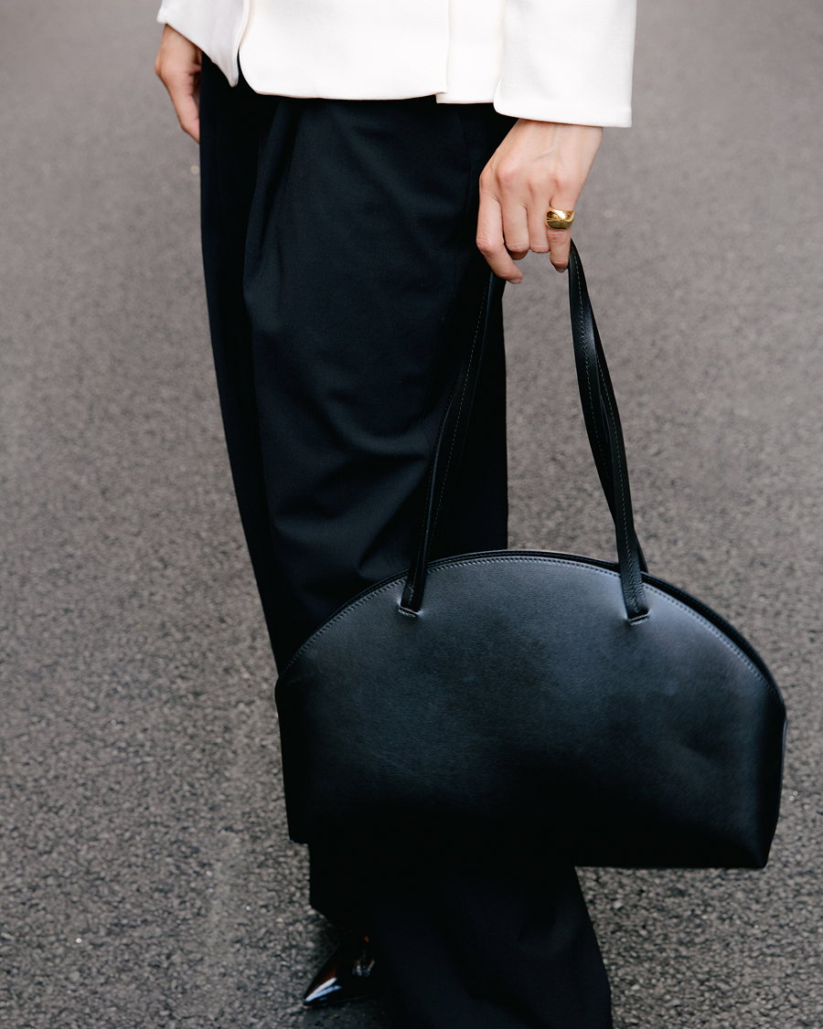 A person in a white jacket and black trousers holding a black, half-moon shaped leather handbag.