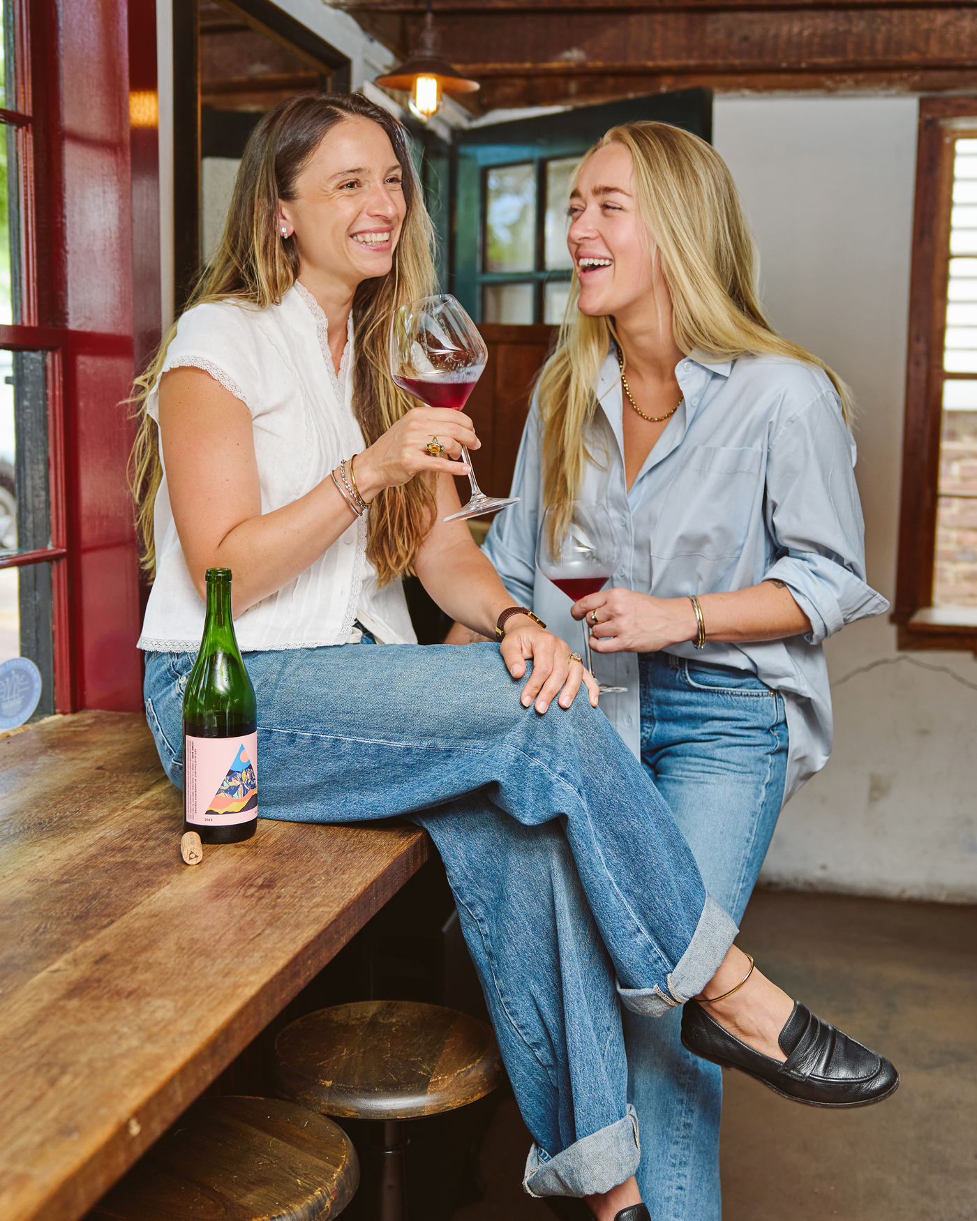 Two women in a rustic setting laugh while drinking red wine together at a wooden bar.