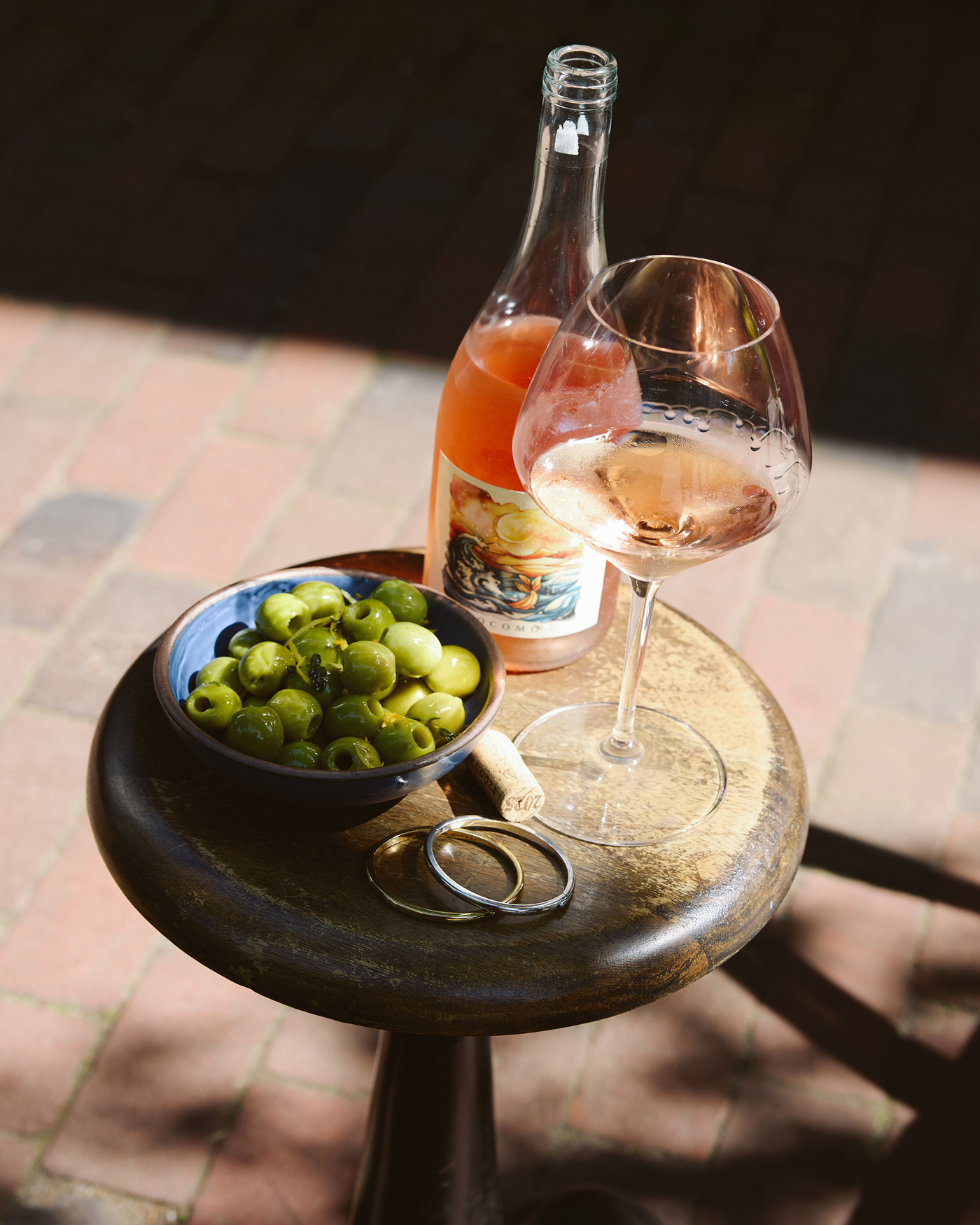 A bottle and glass of rosé wine with a bowl of olives on a wooden stool outdoors.