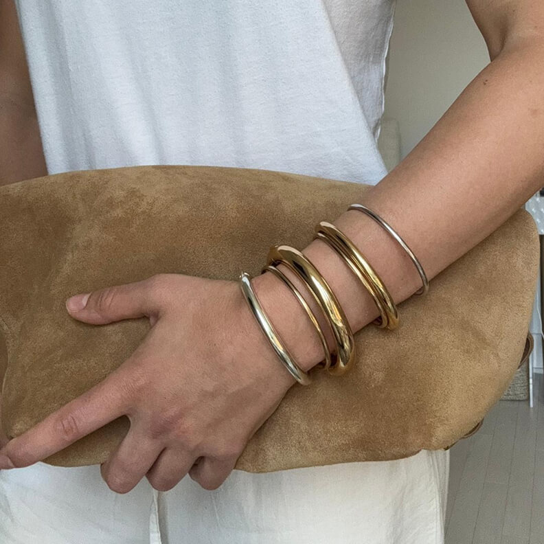 A person wearing a white top holds a brown suede clutch, their arm adorned with gold and silver bangles.