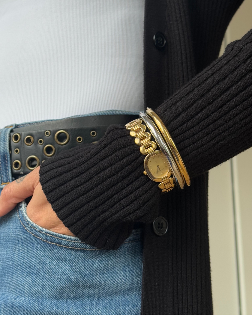 Person wearing a black sweater, gold watch, bracelets, and jeans with a studded belt.