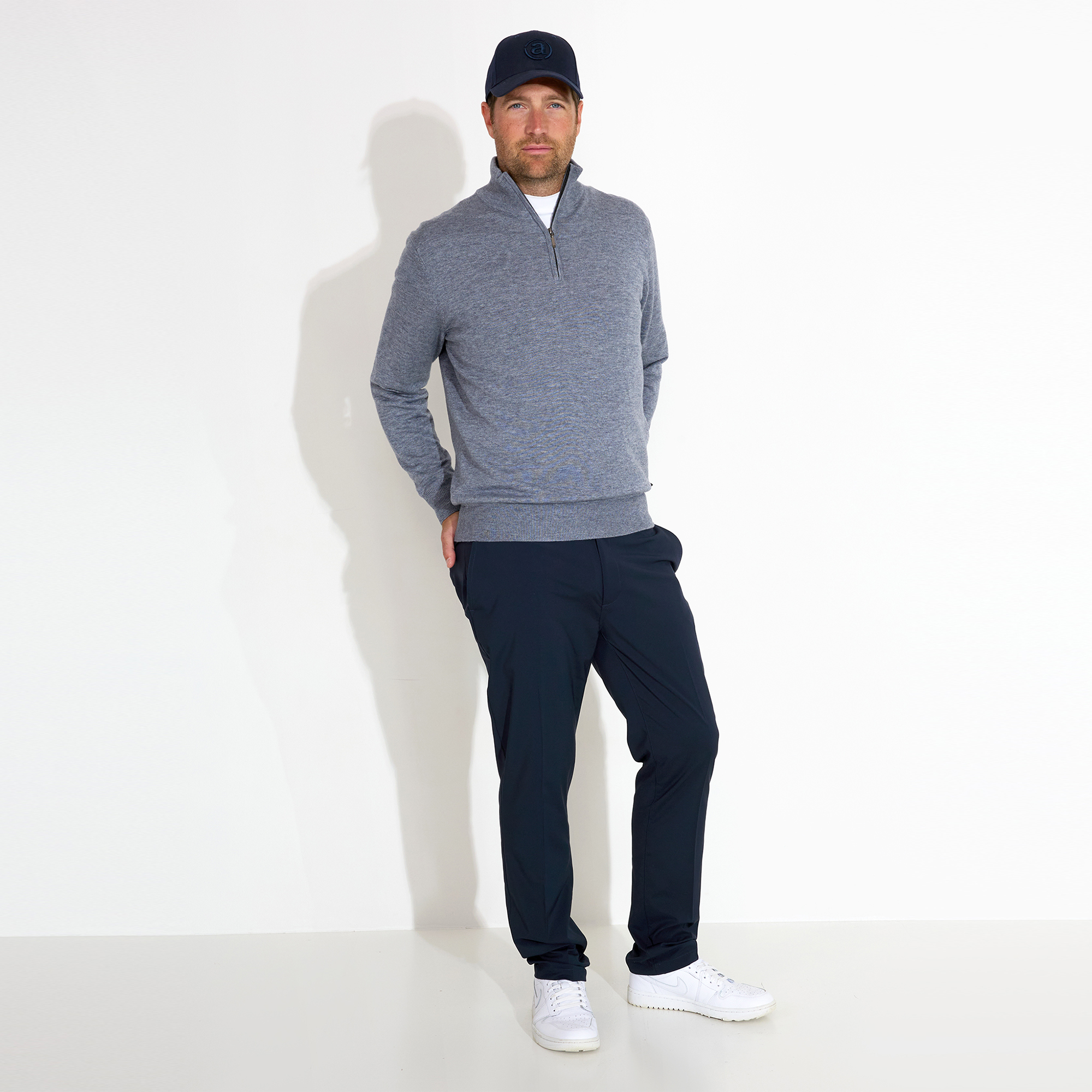 Man in casual outfit with gray sweater and cap, leaning against white background.