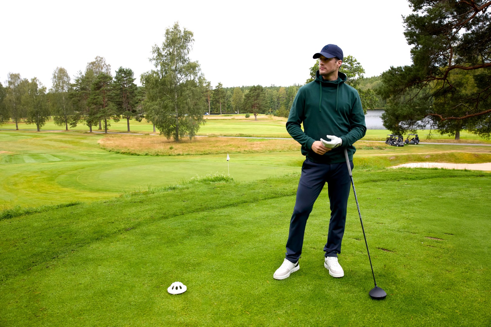 A man in a green hoodie and a blue cap stands on a golf course holding a driver.