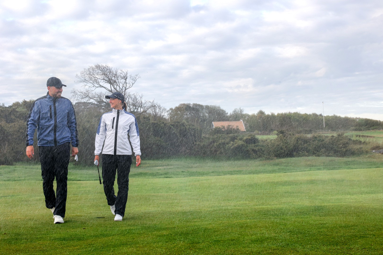 A man and a woman in rain jackets walk across a golf course in the rain.
