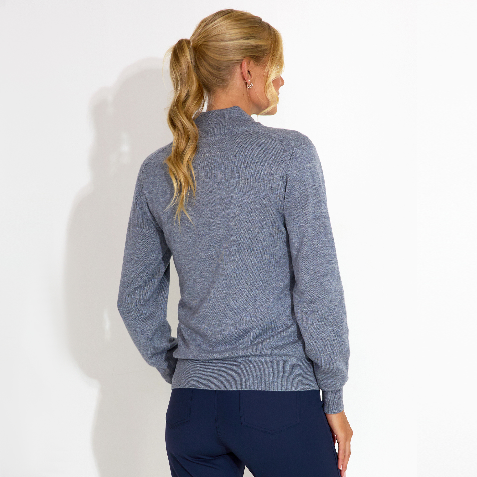 Person with long hair in ponytail, wearing a gray sweater and dark pants, standing against a white background.