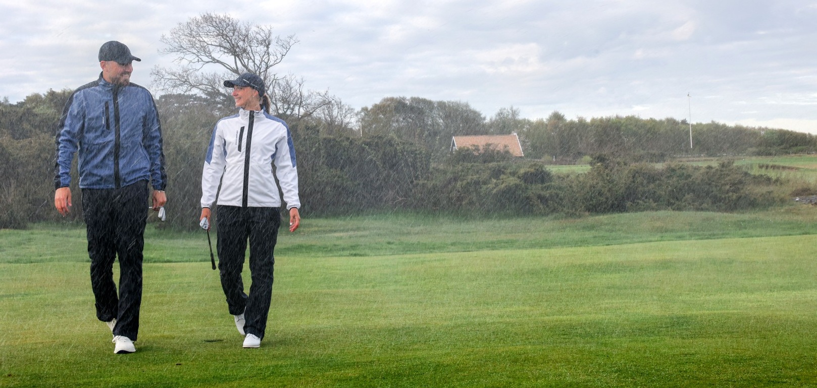 A man and a woman in rain jackets walk across a golf course in the rain.