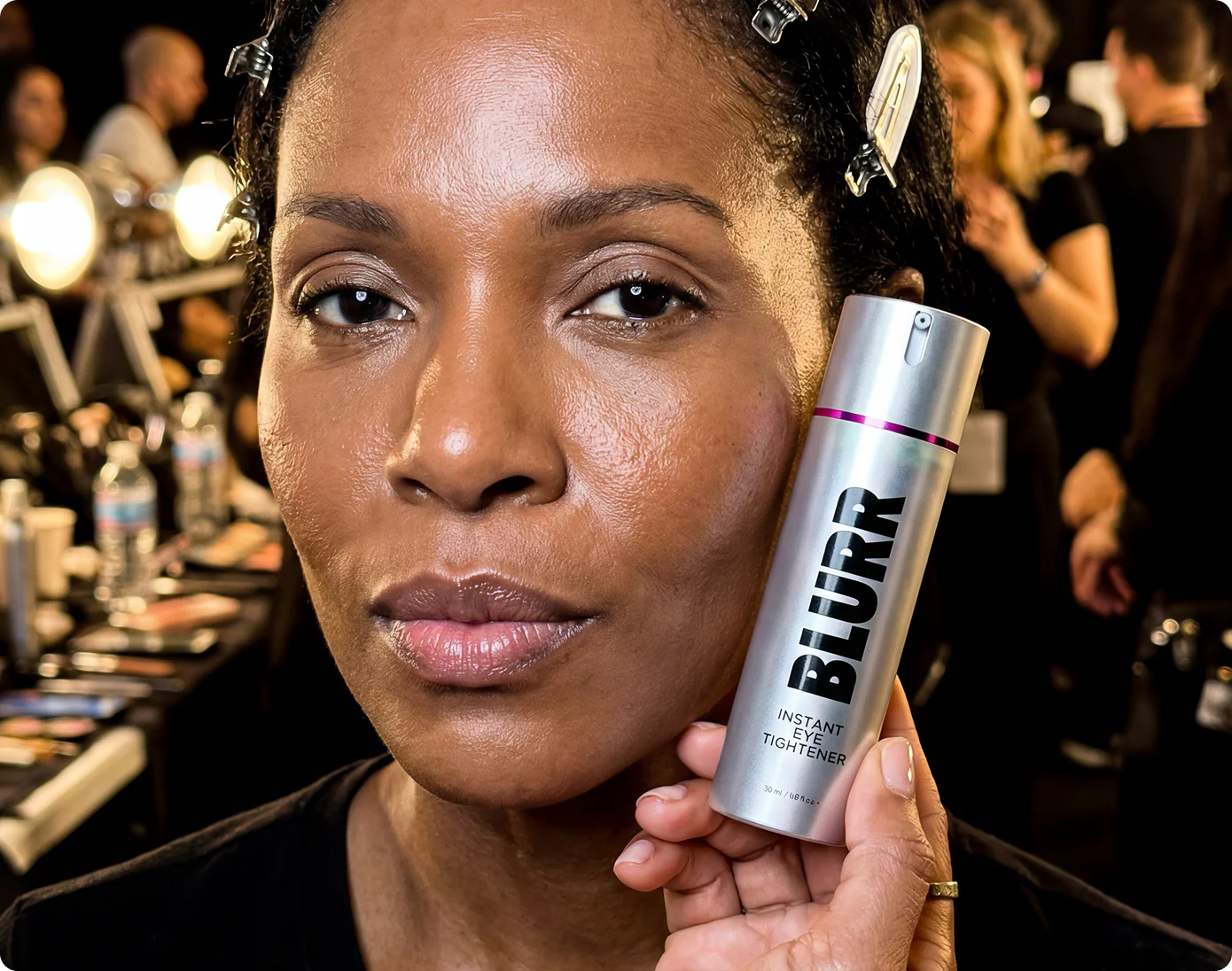 A woman holds a silver bottle of Blurr Instant Eye Tightener next to her face backstage.