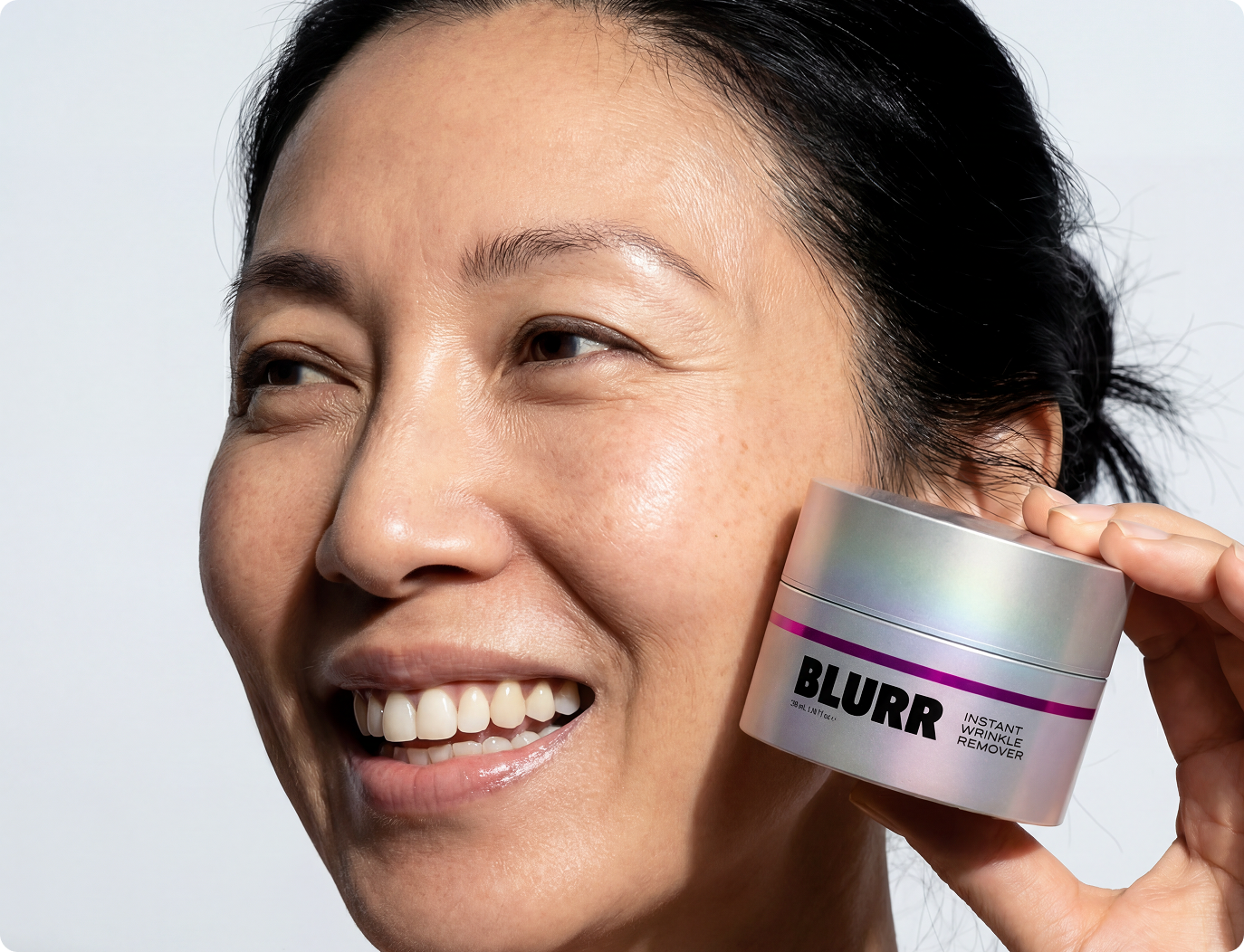 A smiling woman holds a silver jar of Blurr Instant Wrinkle Remover next to her cheek.