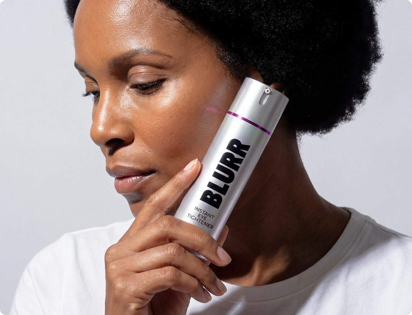 A Black woman holds a silver bottle of 'Blurr' Instant Eye Tightener to her cheek.