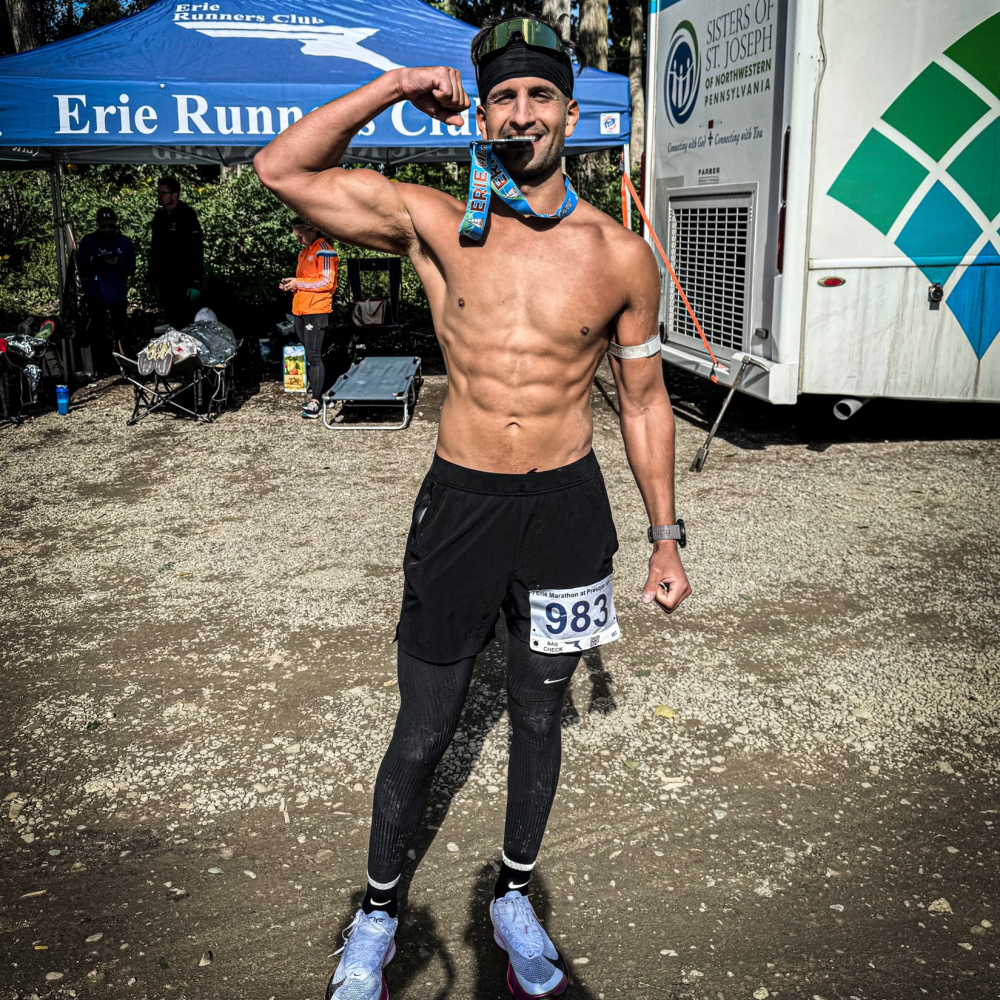 Man flexing with a medal, wearing running gear, under a tent.