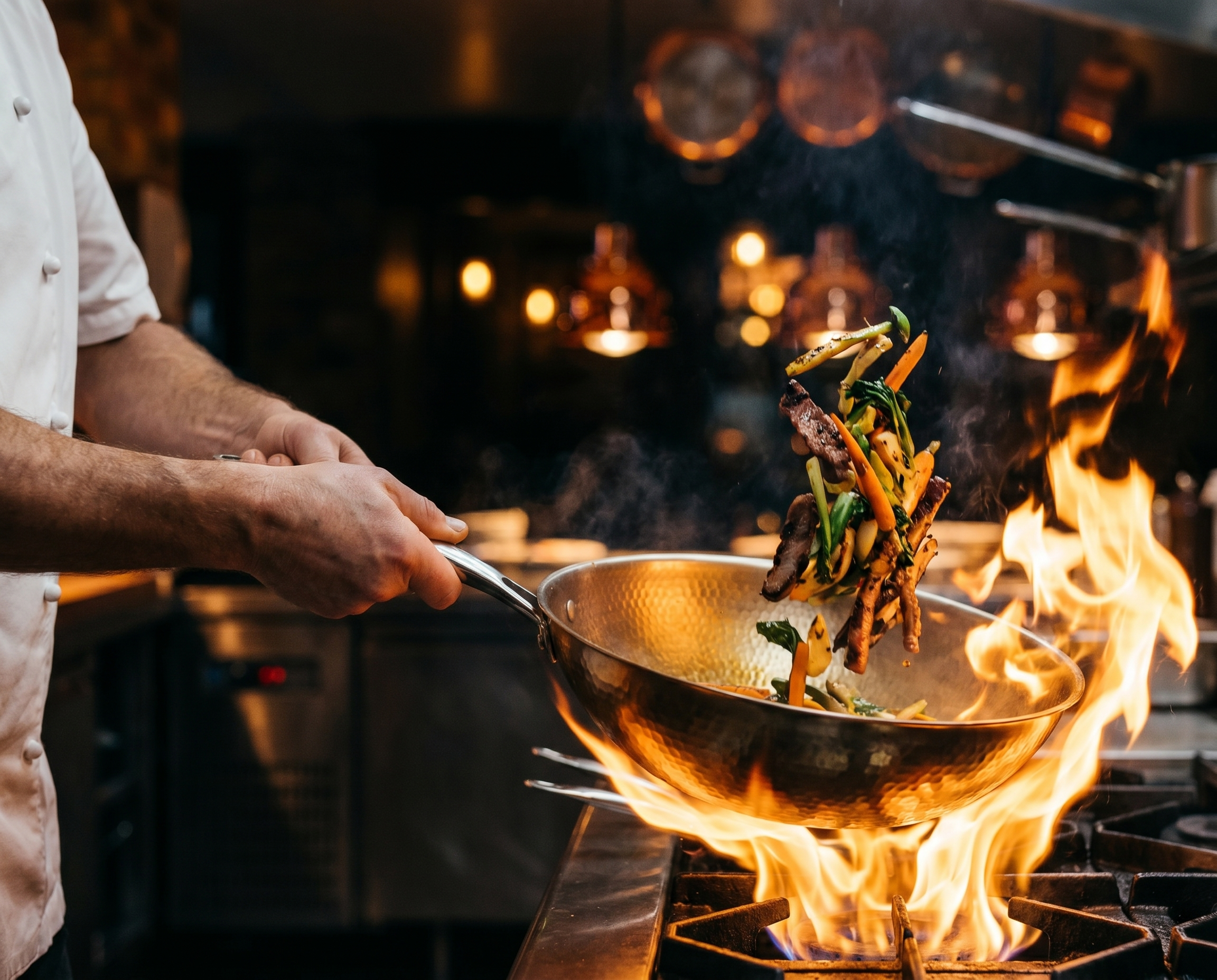 A chef tossing meat and vegetables in a pan over a large open flame in a kitchen.