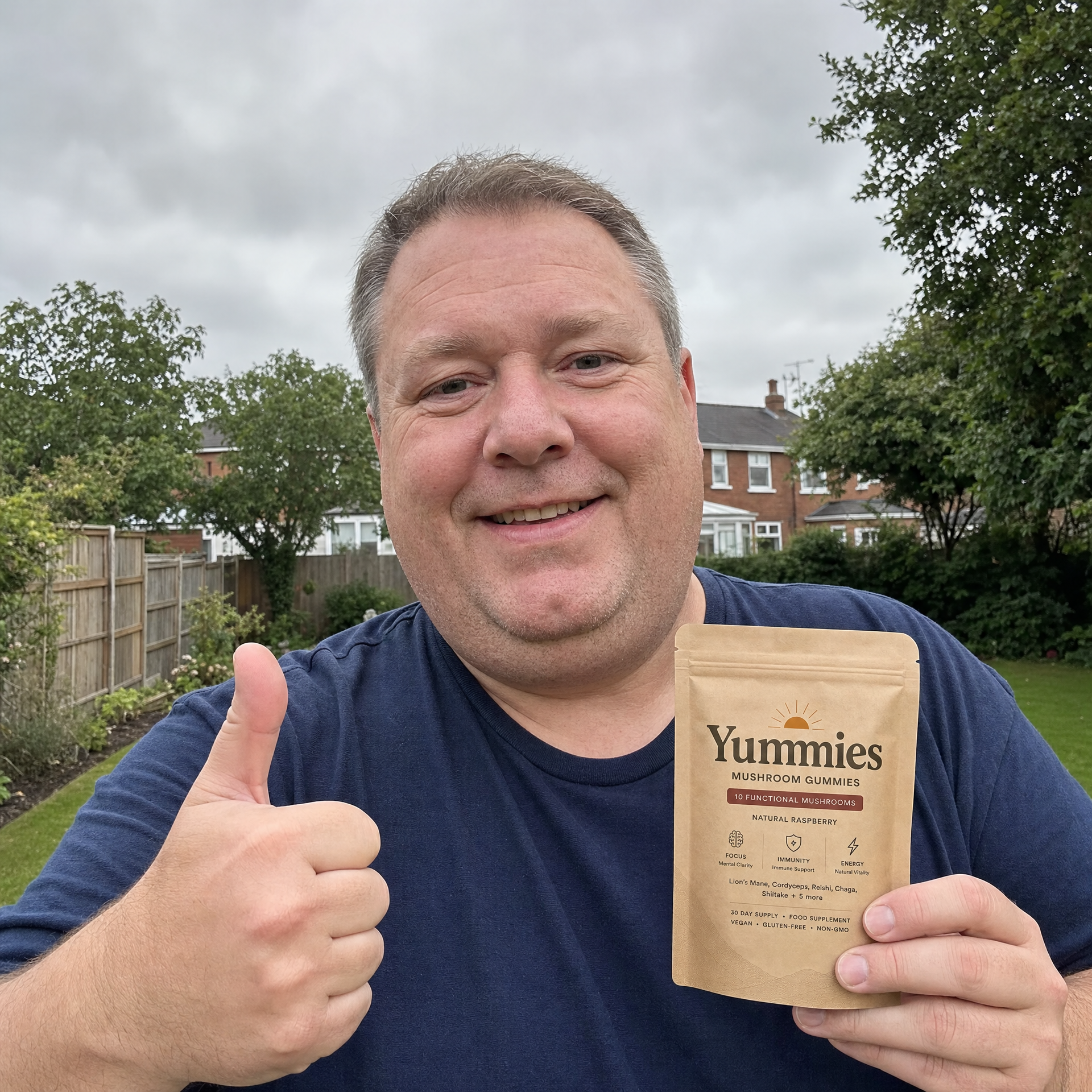 A smiling man holds a pouch of Yummies Mushroom Gummies and gives a thumbs-up in a backyard.
