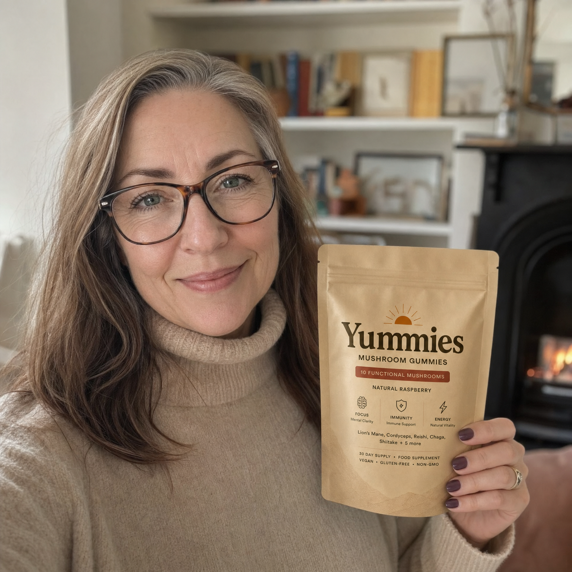 A smiling woman in glasses and a beige turtleneck holds a pouch of Yummies Mushroom Gummies.