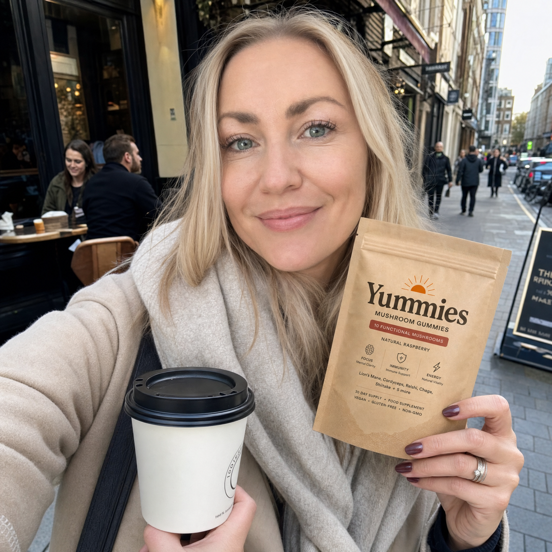 A smiling woman on a city street holds a coffee cup and a pouch of Yummies Mushroom Gummies.