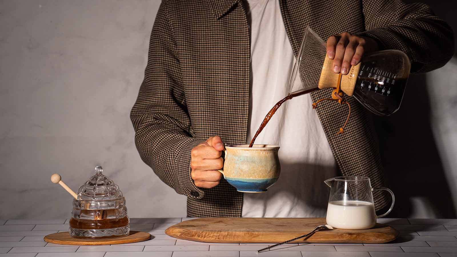 Person pouring coffee into a mug, with honey jar and milk pitcher on table.