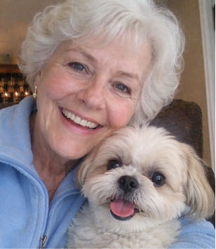 A smiling woman with short white hair holding a small, fluffy, light-colored dog.