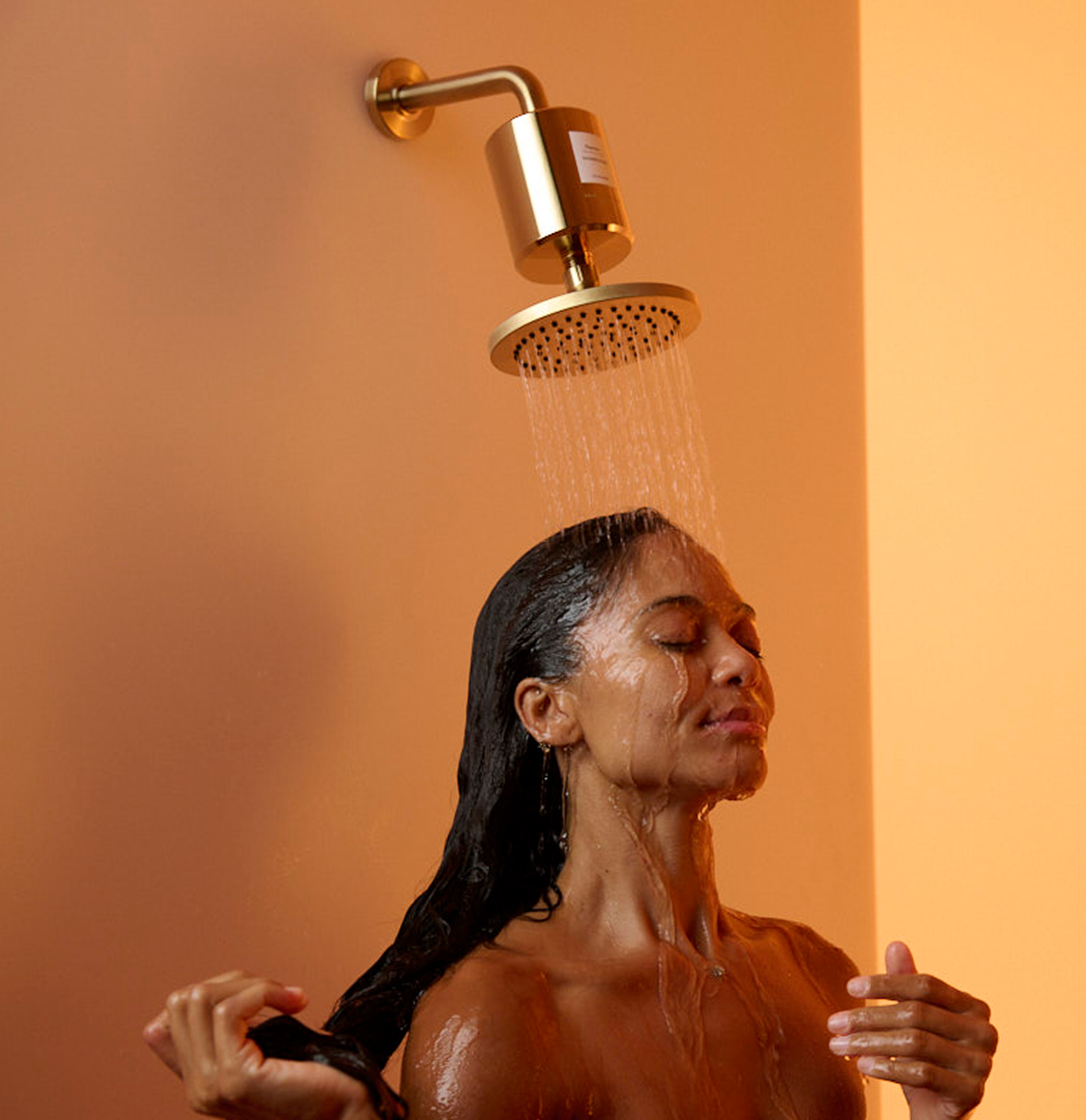 Person enjoying a shower under a gold showerhead with orange background.