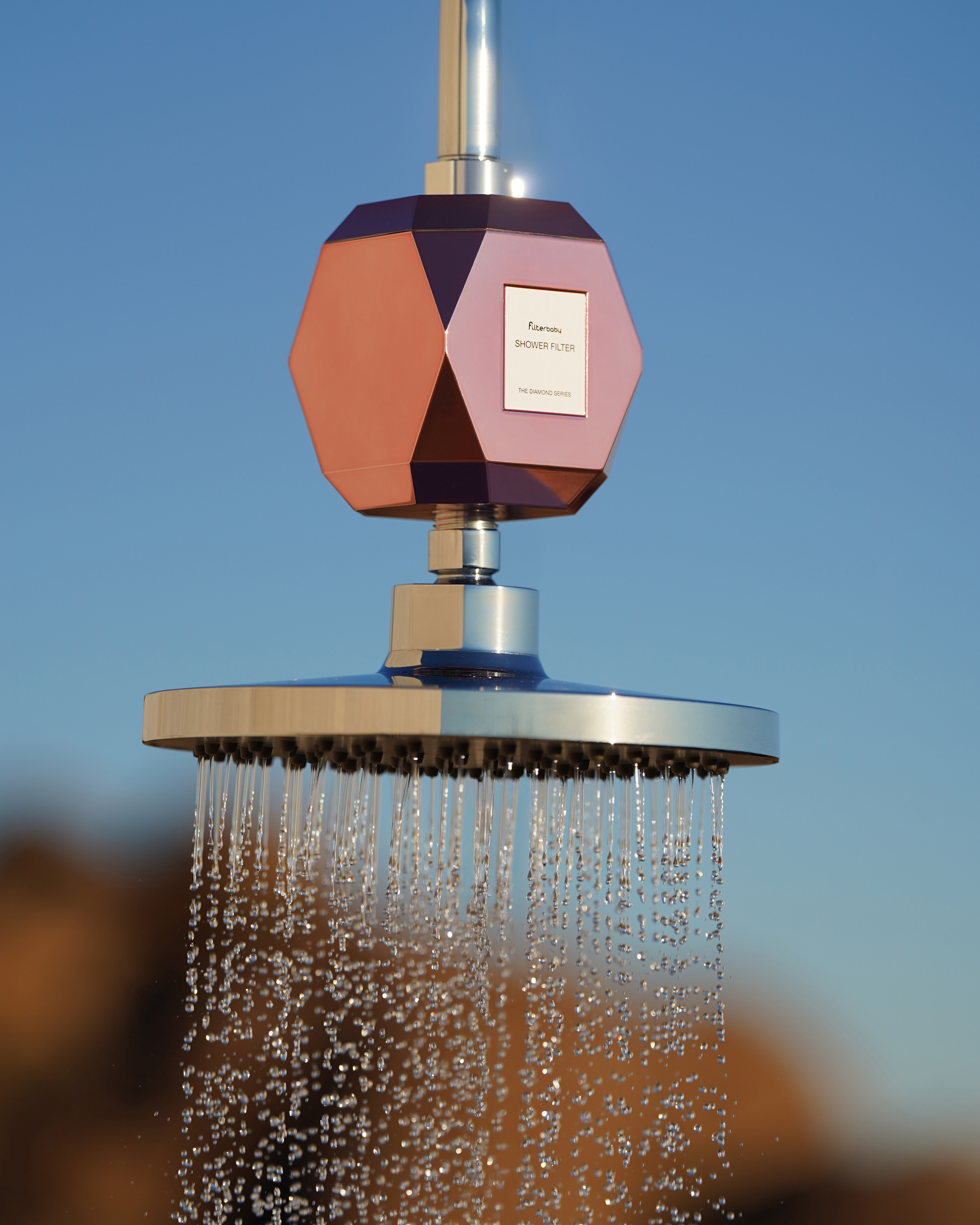 Shower head with a geometric filter, water flowing, against a clear blue sky.