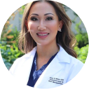 Person in a white coat smiling outdoors with greenery in the background.
