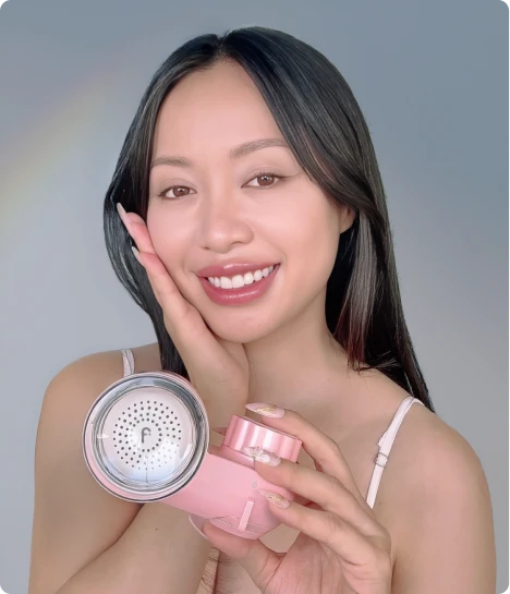 A smiling woman with dark hair holds a pink and silver facial device while touching her cheek.