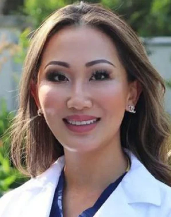 A close-up headshot of a smiling woman with long brown hair, wearing a white coat.