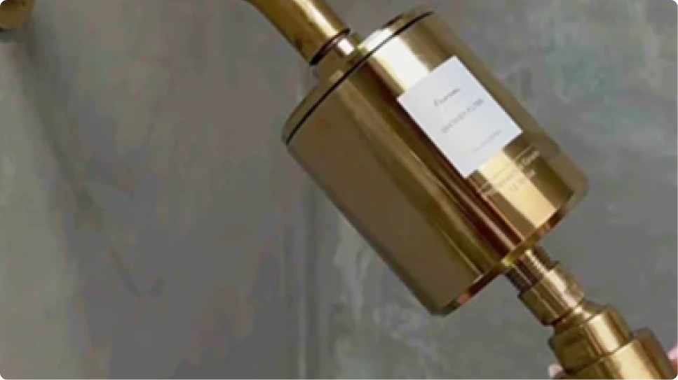A close-up of a gold-colored shower filter attached to a shower arm against a gray wall.