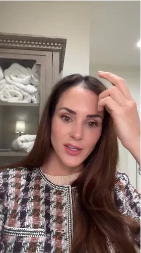 A woman with long brown hair wearing a patterned jacket touches her forehead while looking forward.