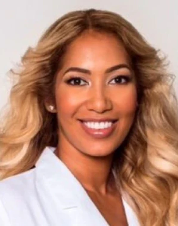 A headshot of a smiling woman with long, blonde, wavy hair wearing a white coat.
