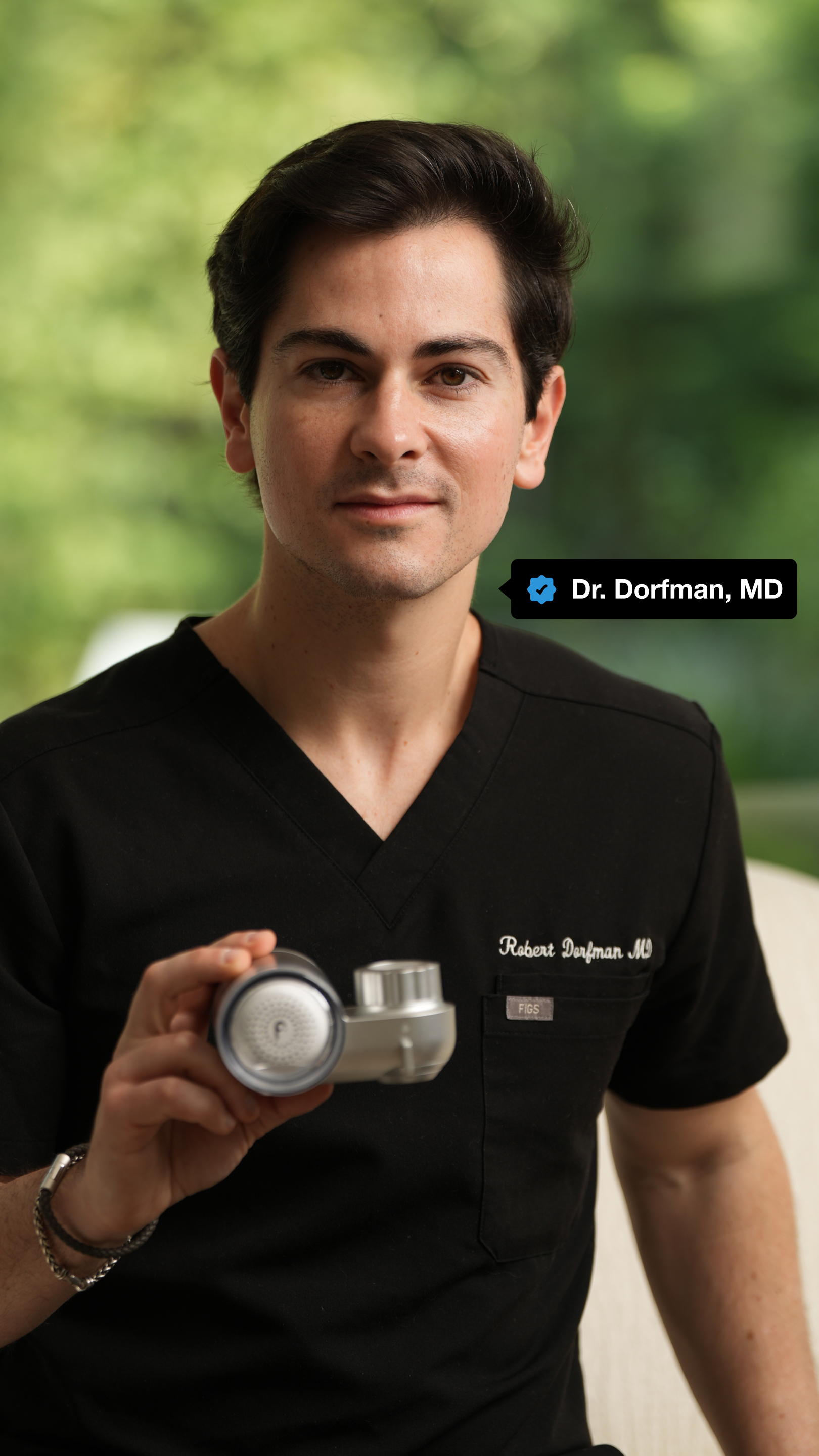 Person in black scrubs holding a medical device, with a blurred green background.