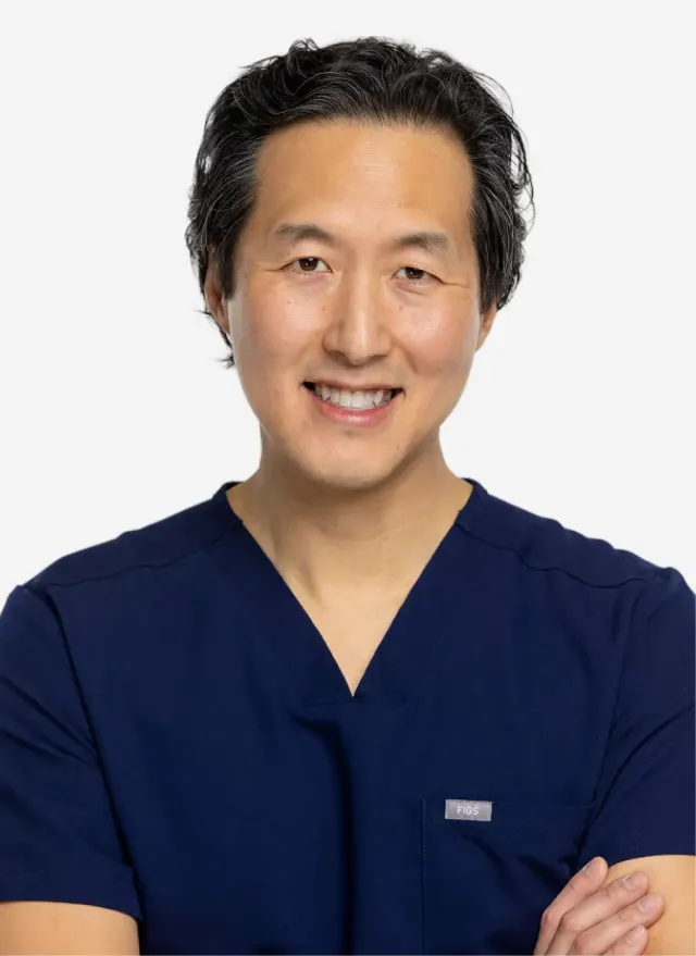 A smiling middle-aged East Asian man with dark hair, wearing navy blue scrubs against a white background.