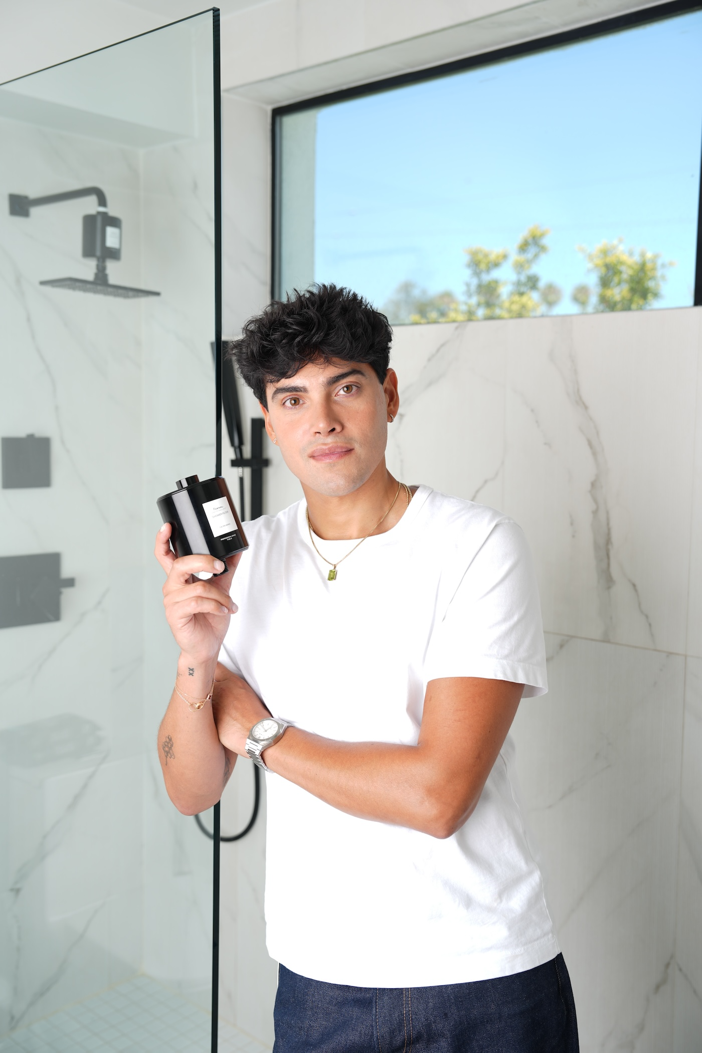 A man in a white t-shirt and jeans stands in a modern bathroom holding a black bottle.
