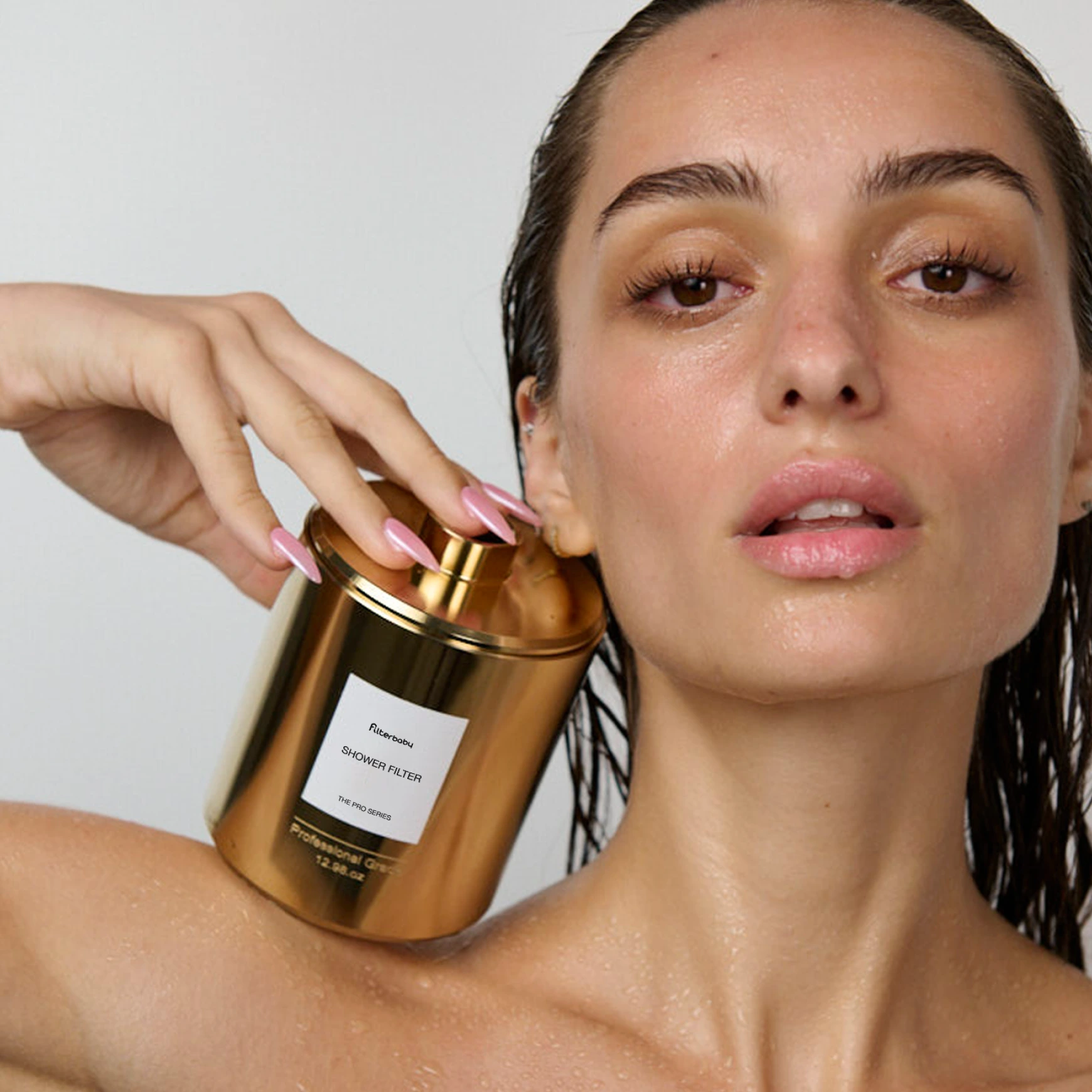 A woman with wet skin and hair holds a gold Filterbaby shower filter against her shoulder.