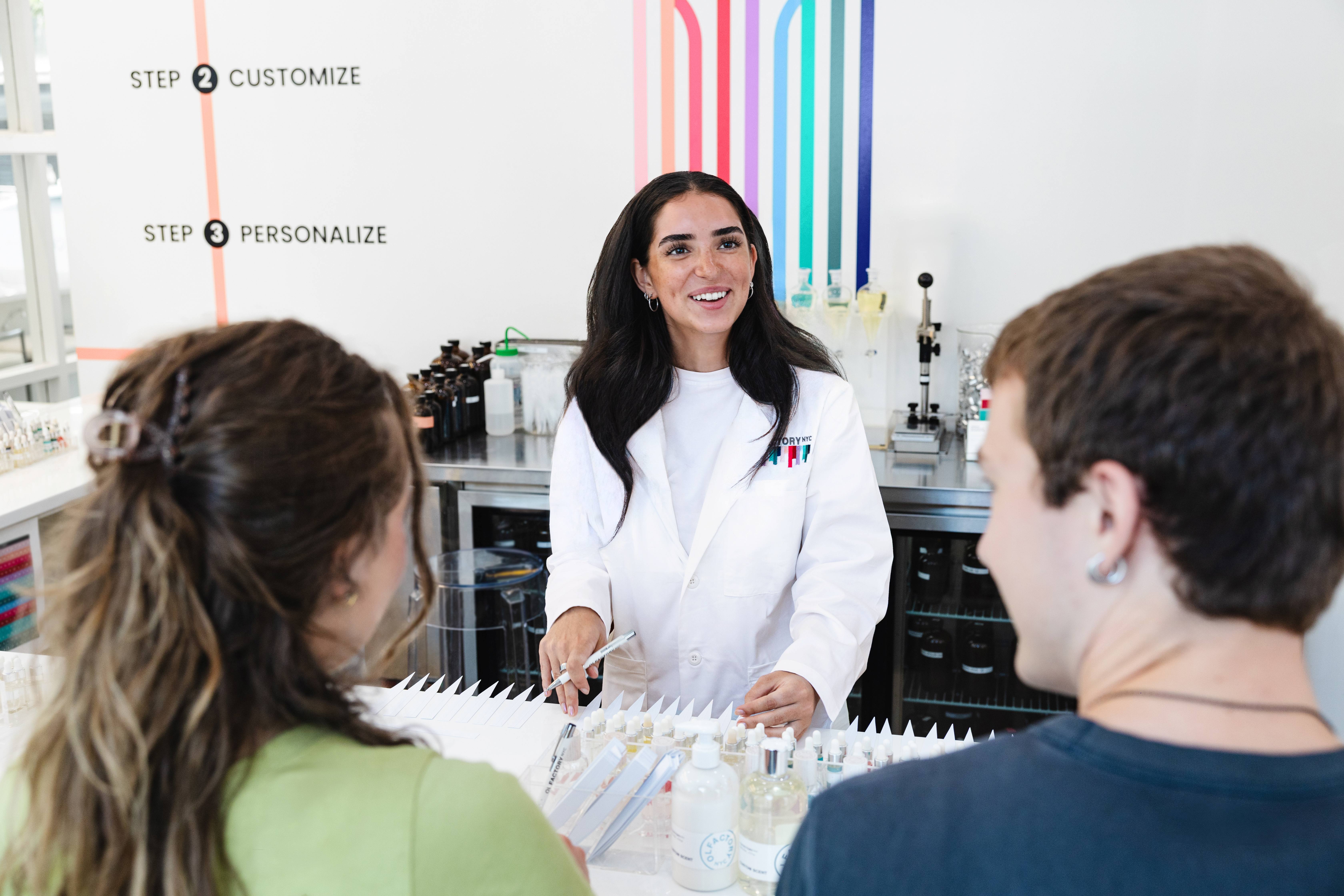 A woman in a lab coat smiles while talking to two people at a fragrance creation counter.