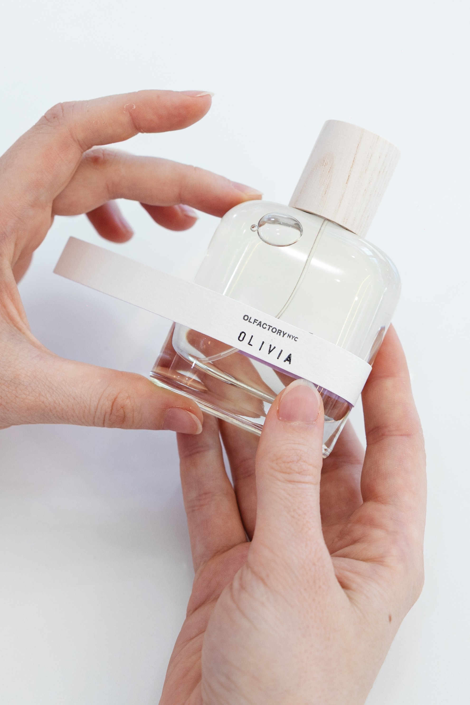 Hands holding a perfume bottle labeled 'Olivia' with a wooden cap.