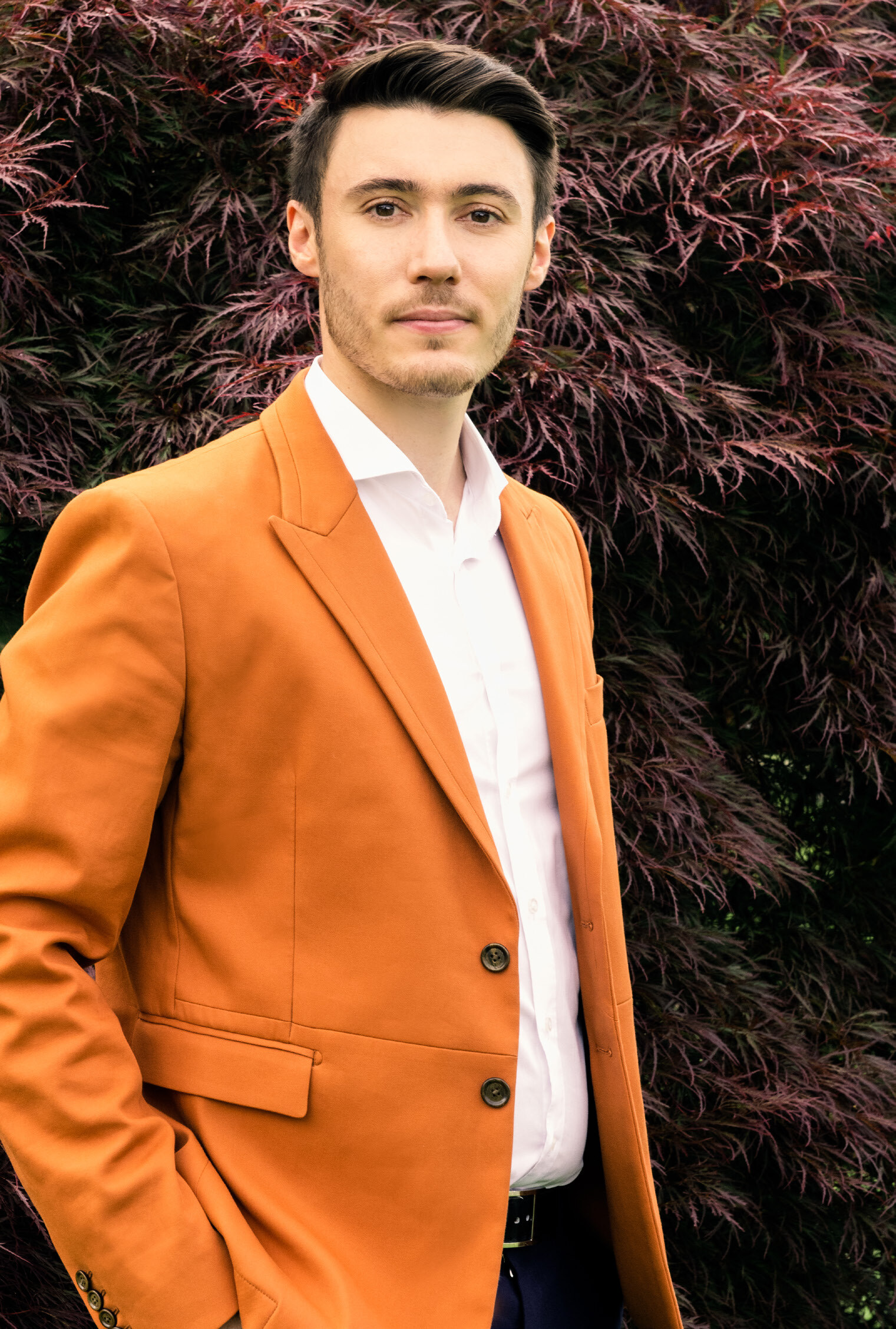 A man in a bright orange blazer over a white shirt stands in front of a dark, leafy bush.